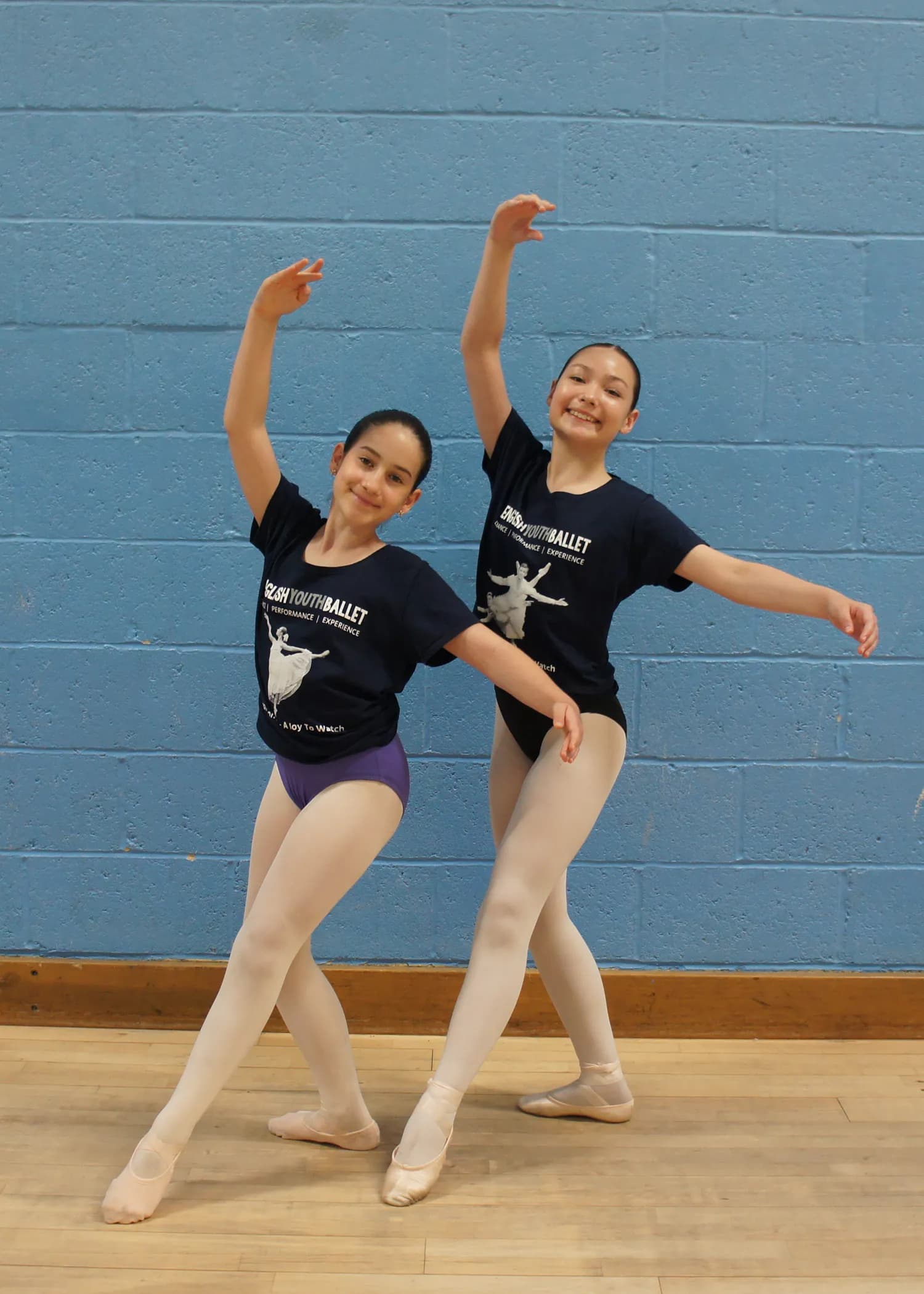 Two young ballerinas wearing dark blue t-shirts, light-colored tights, and ballet shoes pose in a studio with a blue brick wall and wooden floor.